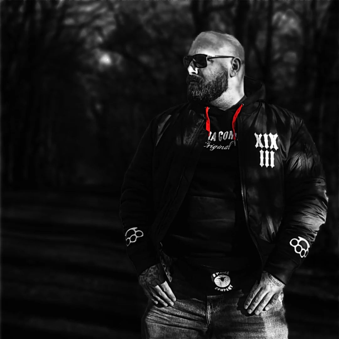 Bärtiger Mann mit Sonnenbrille trägt schwarze Stigma-Company-Jacke und Hoodie in düsterer Schwarz-Weiß-Szene