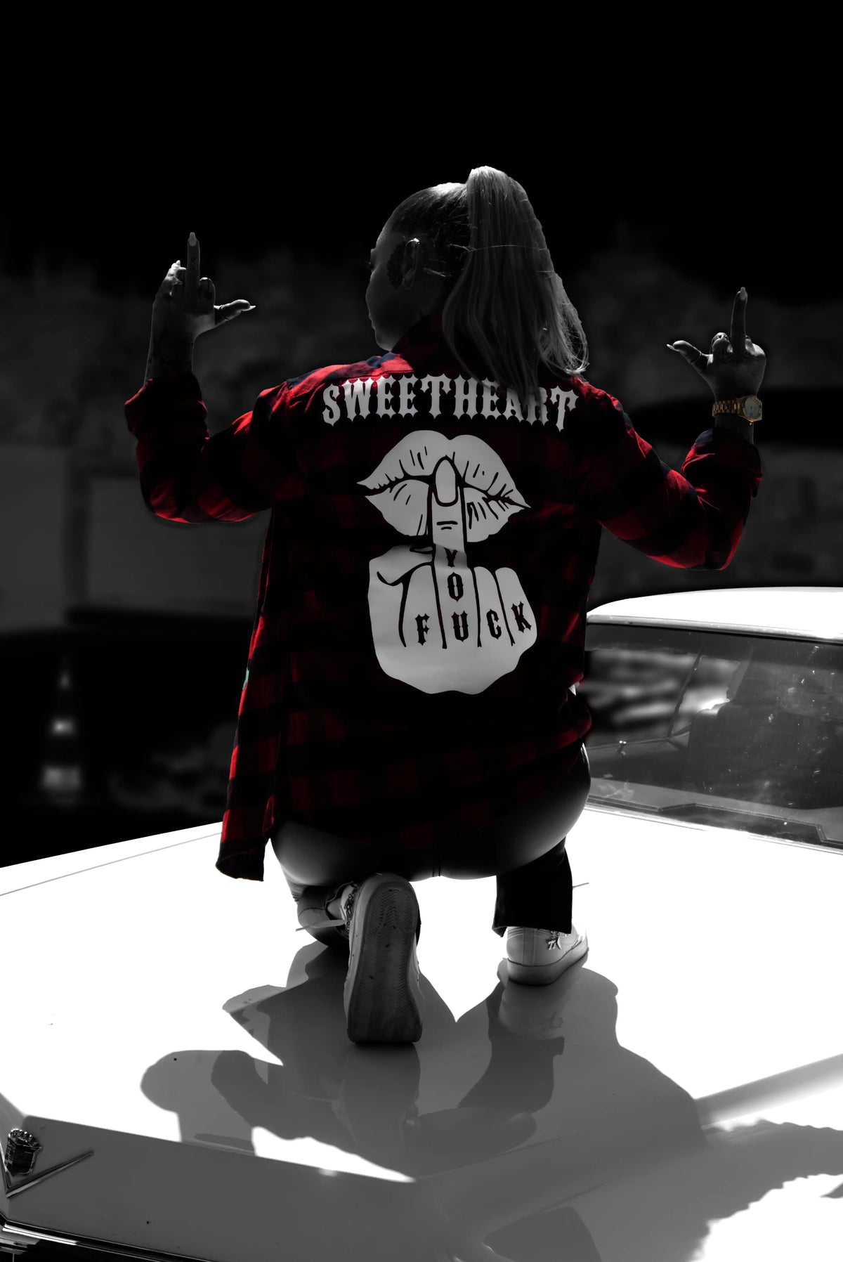 Frau kniet auf einem Cadillac bei Nacht und trägt einen Hemd mit „Sweetheart Fuck You“-Rückenprint, beide Mittelfinger erhoben