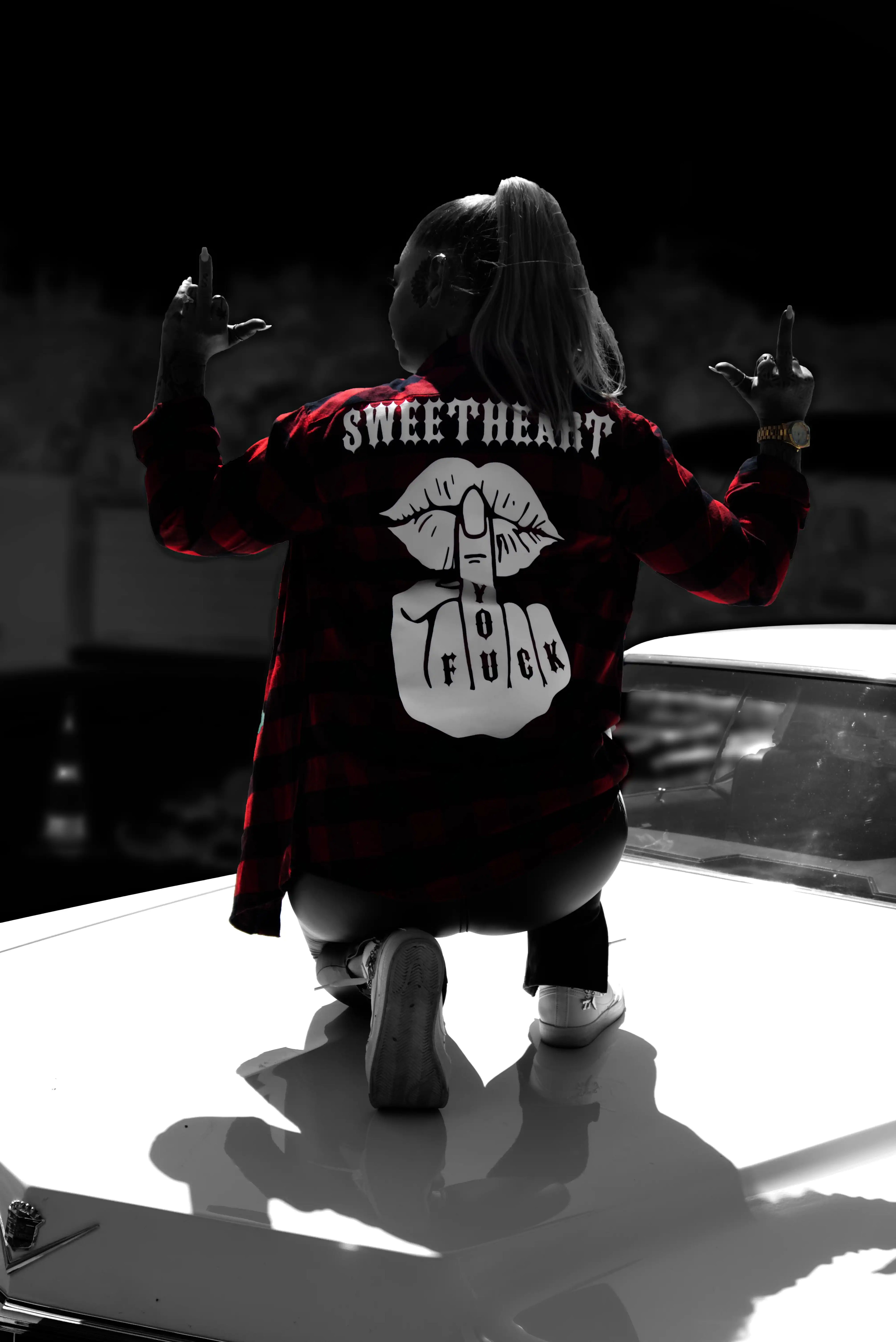 Frau kniet auf einem Cadillac bei Nacht und trägt einen Hemd mit „Sweetheart Fuck You“-Rückenprint, beide Mittelfinger erhoben
