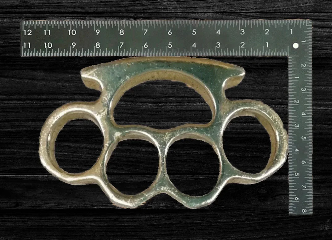 Metall-Schlagring mit vier Fingeröffnungen, daneben ein Lineal zur Größenangabe auf dunklem Hintergrund